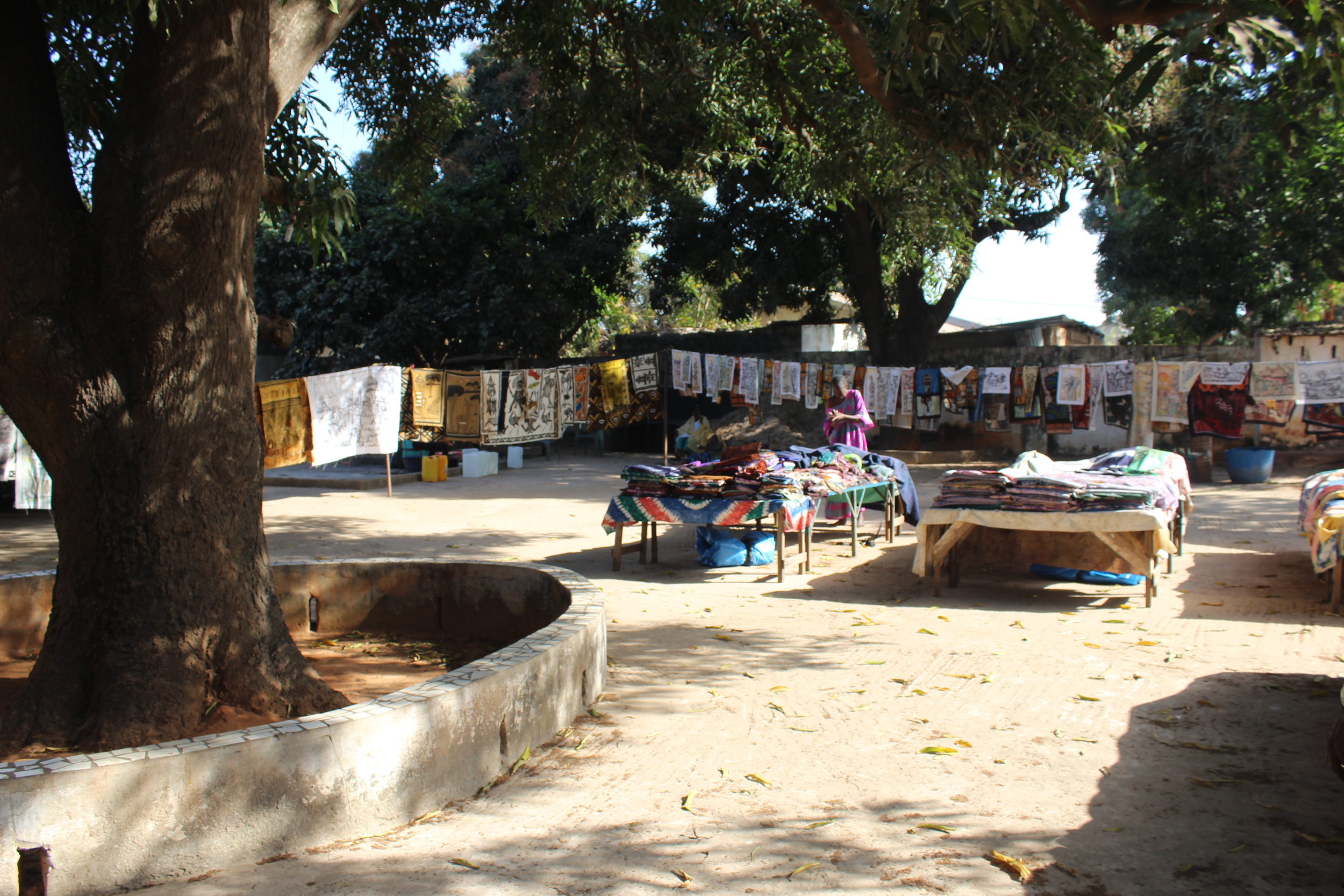 Artwork hung out to dry under a large Mango tree. The central tables contain hundreds of folded and ironed fabric or handmade finished items such as tablecloths and bedding sets.